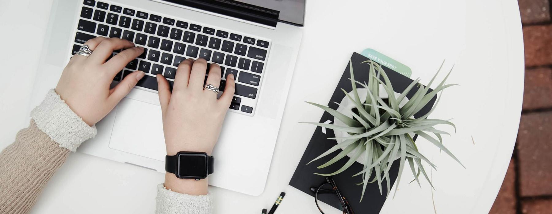 overhead shot of woman working on a laptop at a round table with pencils, her cell phone, glasses, notebook and a potted plant