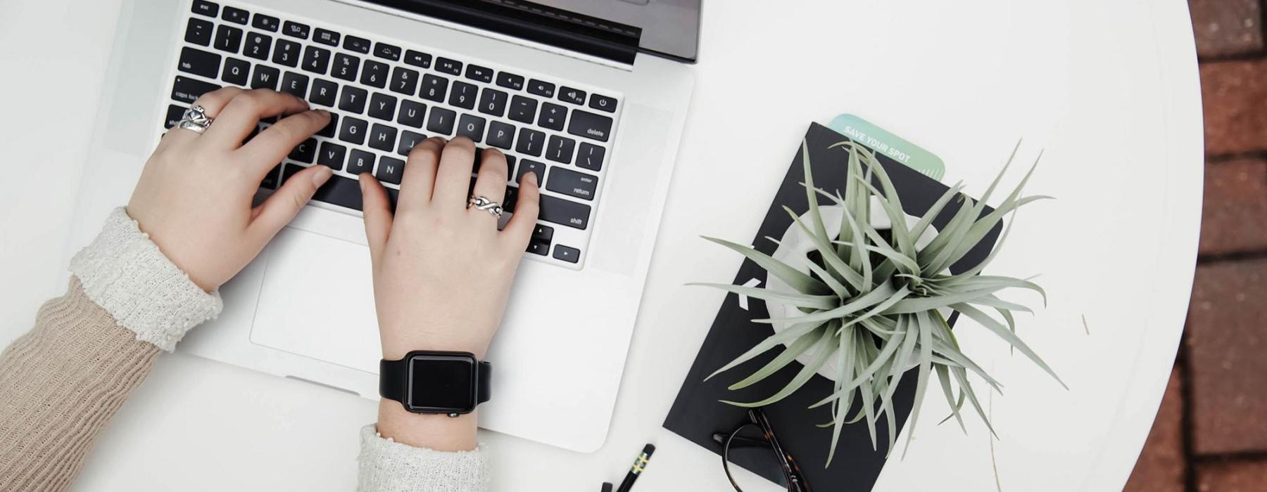 overhead shot of woman working on a laptop at a round table with pencils, her cell phone, glasses, notebook and a potted plant